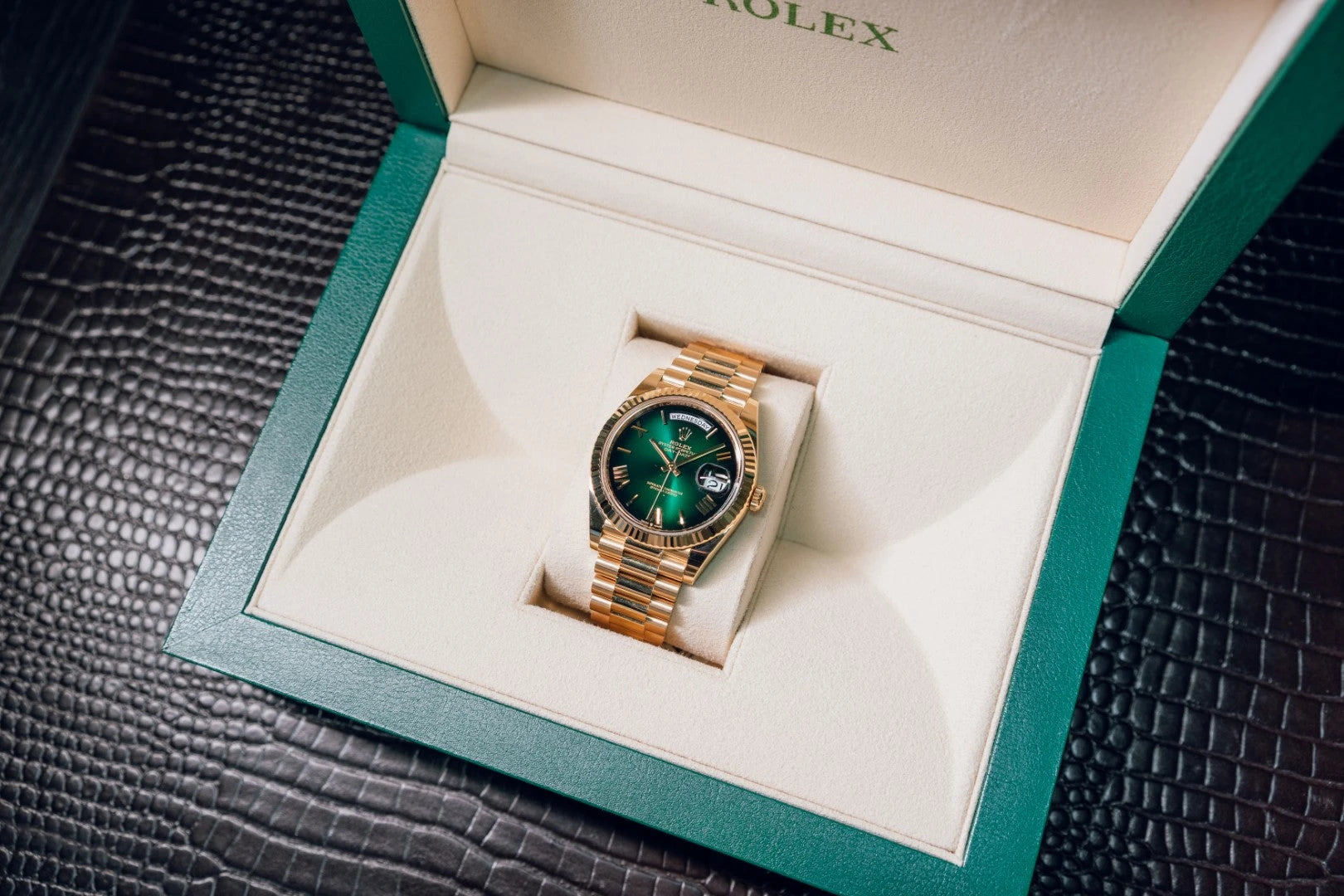 A luxurious gold watch with a green face lies in an open green and cream box on a textured dark surface, suggesting elegance and sophistication.